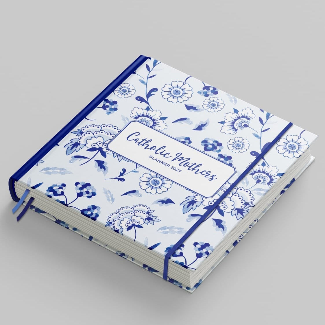 A hardcover journal with a blue and white floral pattern, tied with a matching blue elastic band and featuring a white label that reads 'Catholic Mothers'.
